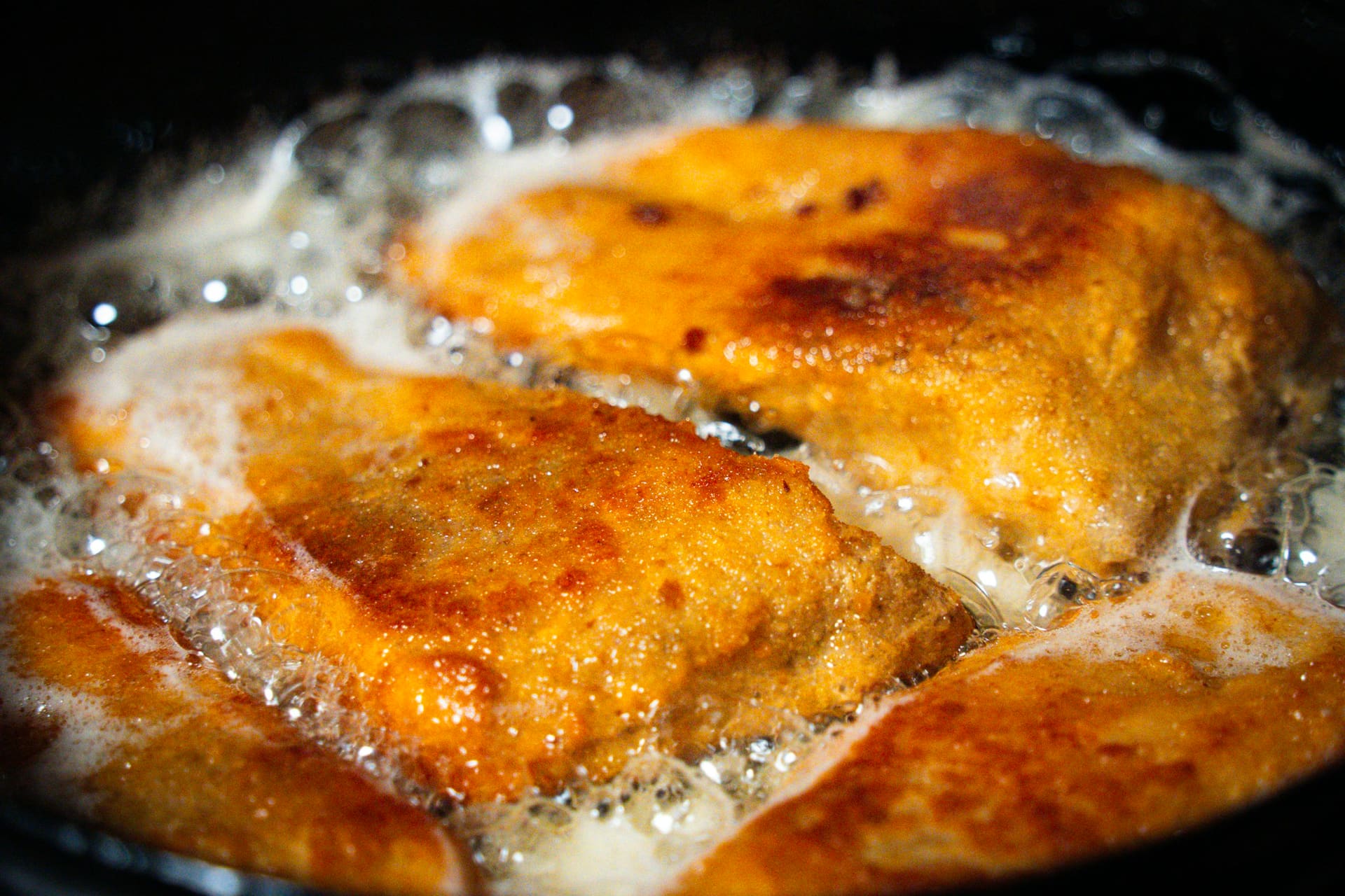 Piezas empanizadas dorándose en aceite caliente, en primer plano sobre fondo oscuro.